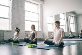 Three pregnant women sitting cross-legged on yoga mats in a bright studio. They appear focused and calm, with natural light streaming in.