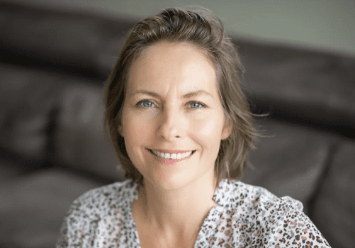 Smiling woman with short hair wearing a floral blouse, sitting indoors on a gray sofa. Warm and inviting atmosphere.