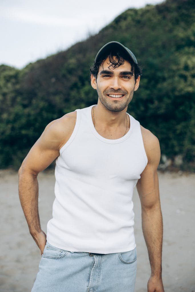 Casually dressed man smiling outdoors on a sandy beach with greenery.