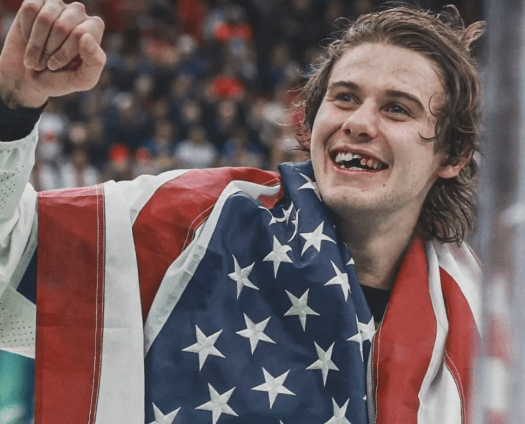 Jack Hughes smiling with a chipped front tooth while celebrating with the American flag draped over his shoulders after a hockey game.
